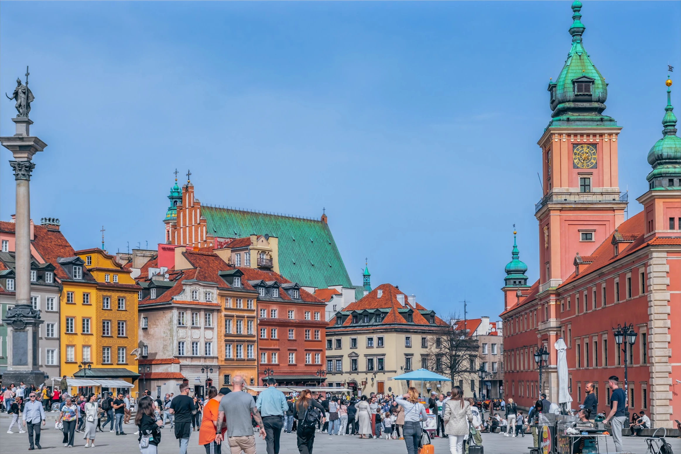 Visitors crossing Warsaw's Castle Square between colourful buildings and the royal castle.
