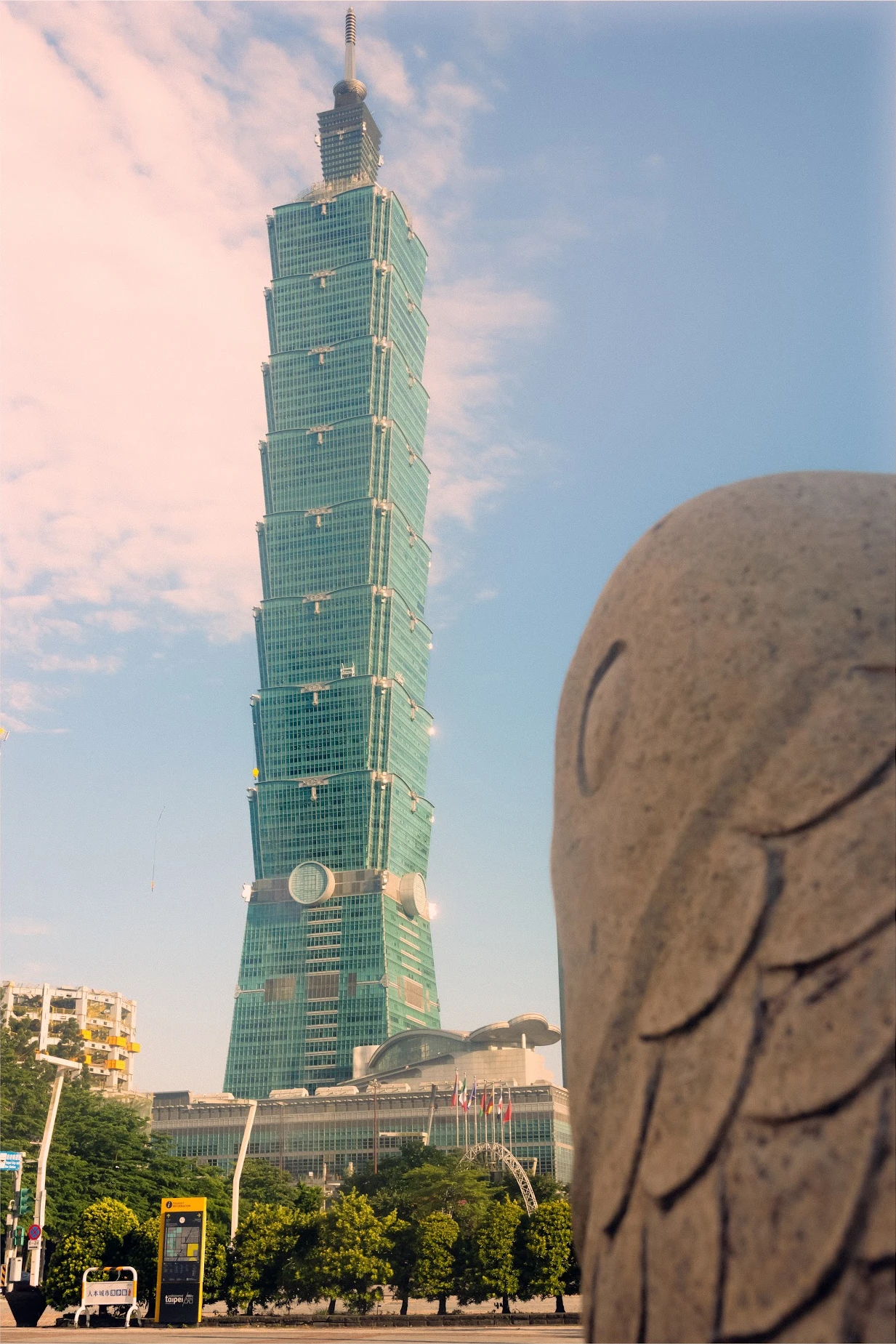 Taipei 101 rising above trees beside a stone sculpture in warm, hazy light.