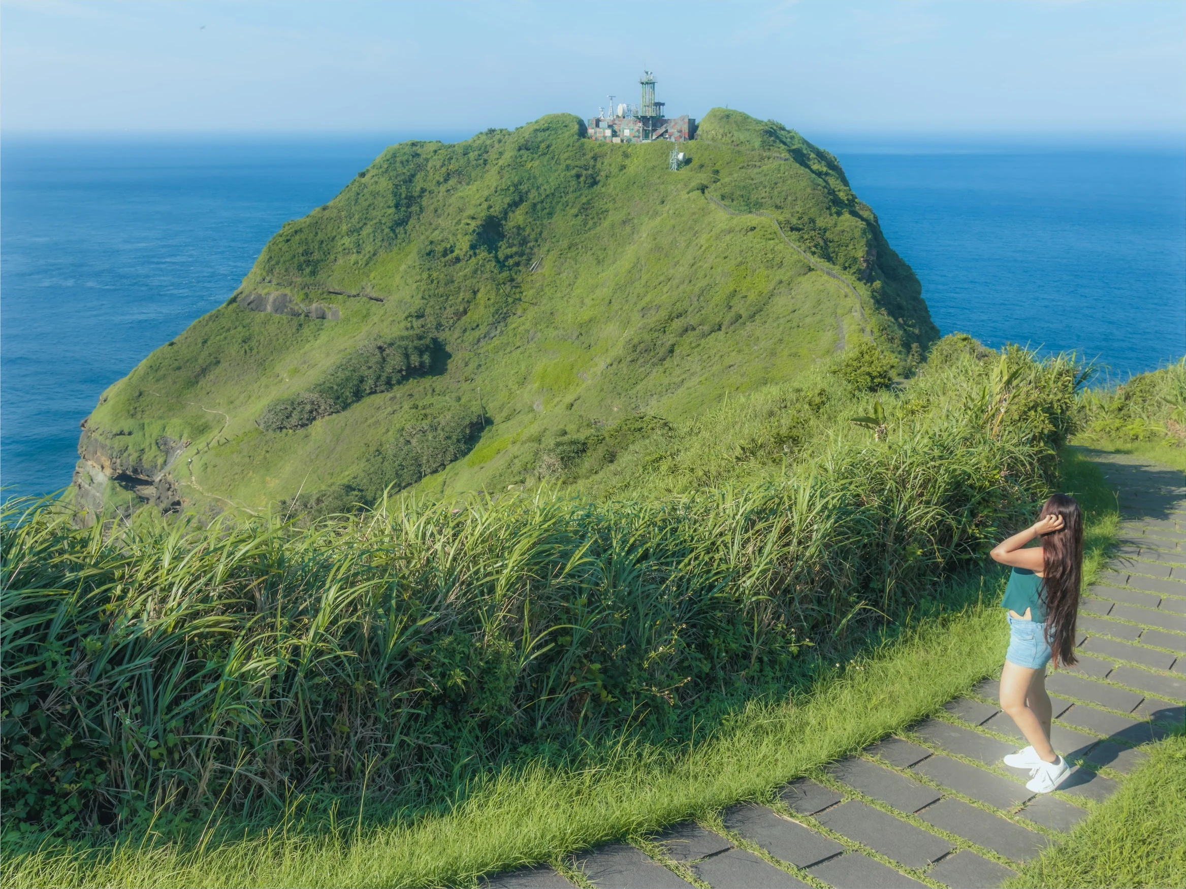A person on stone steps beside a green coastal hill with blue sea beyond.
